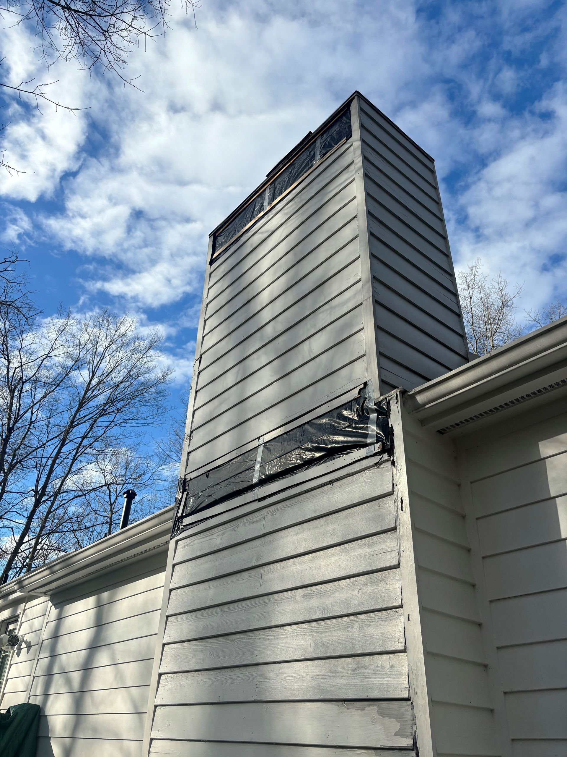 Tall, gray chimney extending from a light-colored house against a cloudy blue sky. Black material covers part of the chimney.