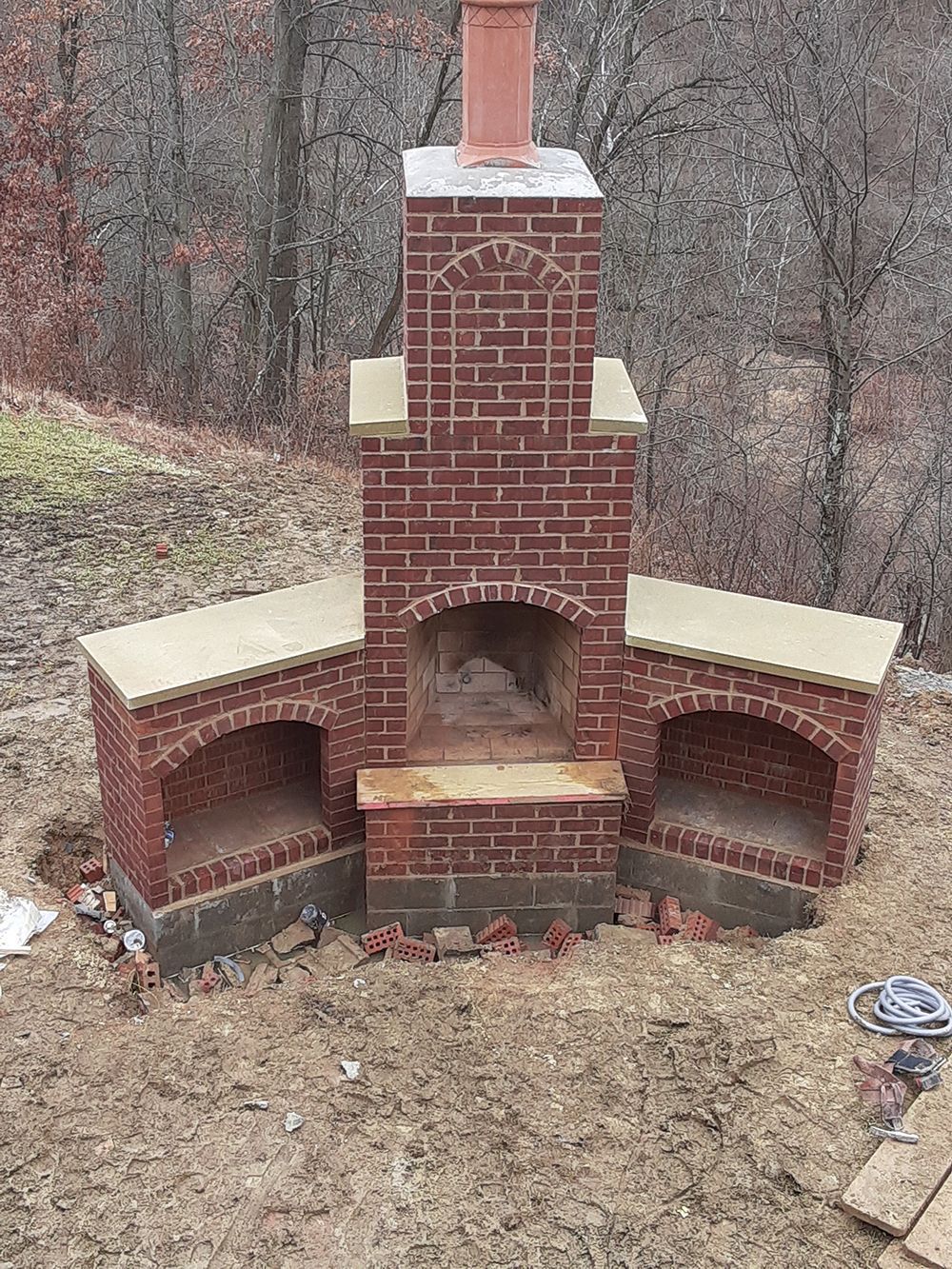 Brick outdoor fireplace with two side compartments, tan accents, and a red chimney in a wooded area.