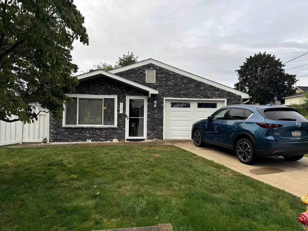 A single-story house with dark siding, a white garage door, and a blue SUV parked in the driveway.