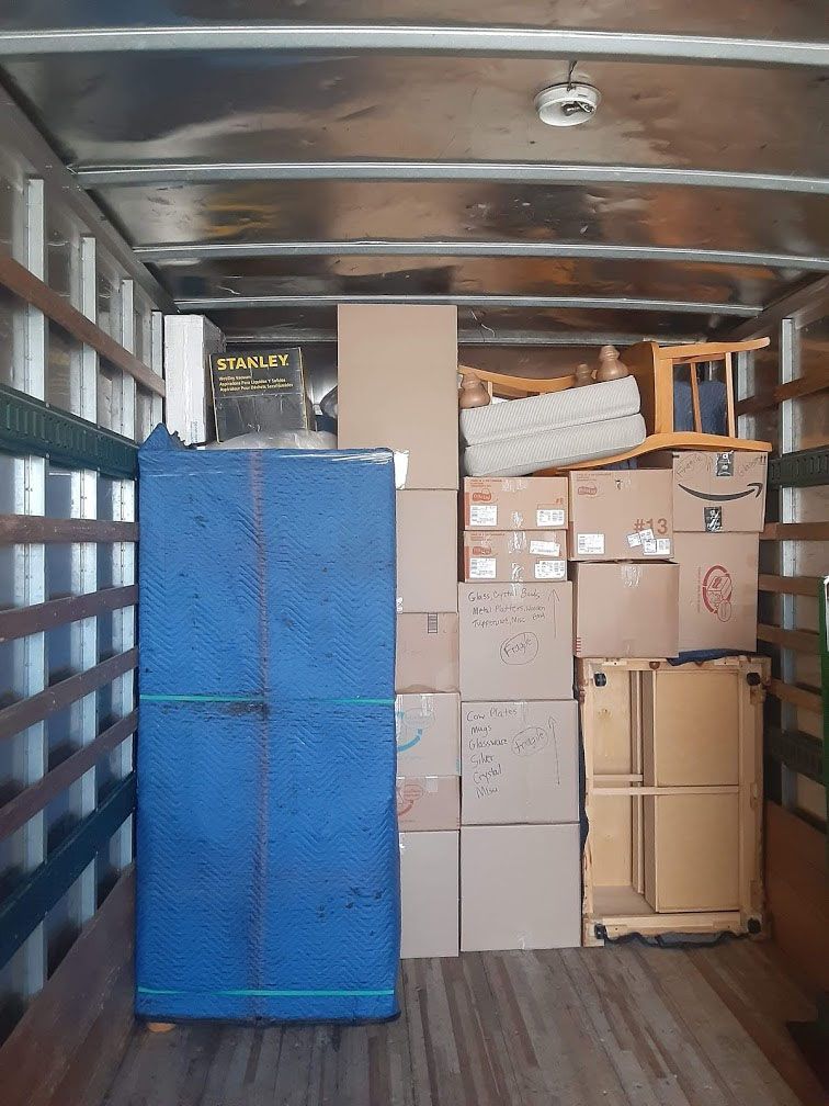 Inside a moving truck packed with furniture and boxes, including a large blue-wrapped item.