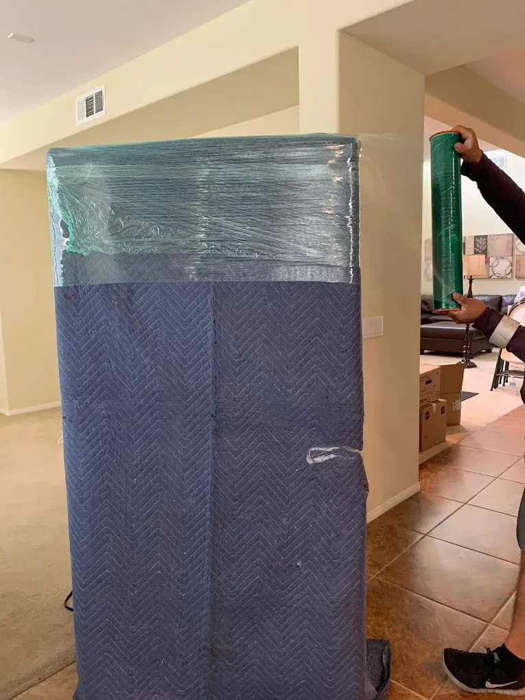 A person wrapping a blue furniture pad-covered item with green plastic wrap, inside a house.