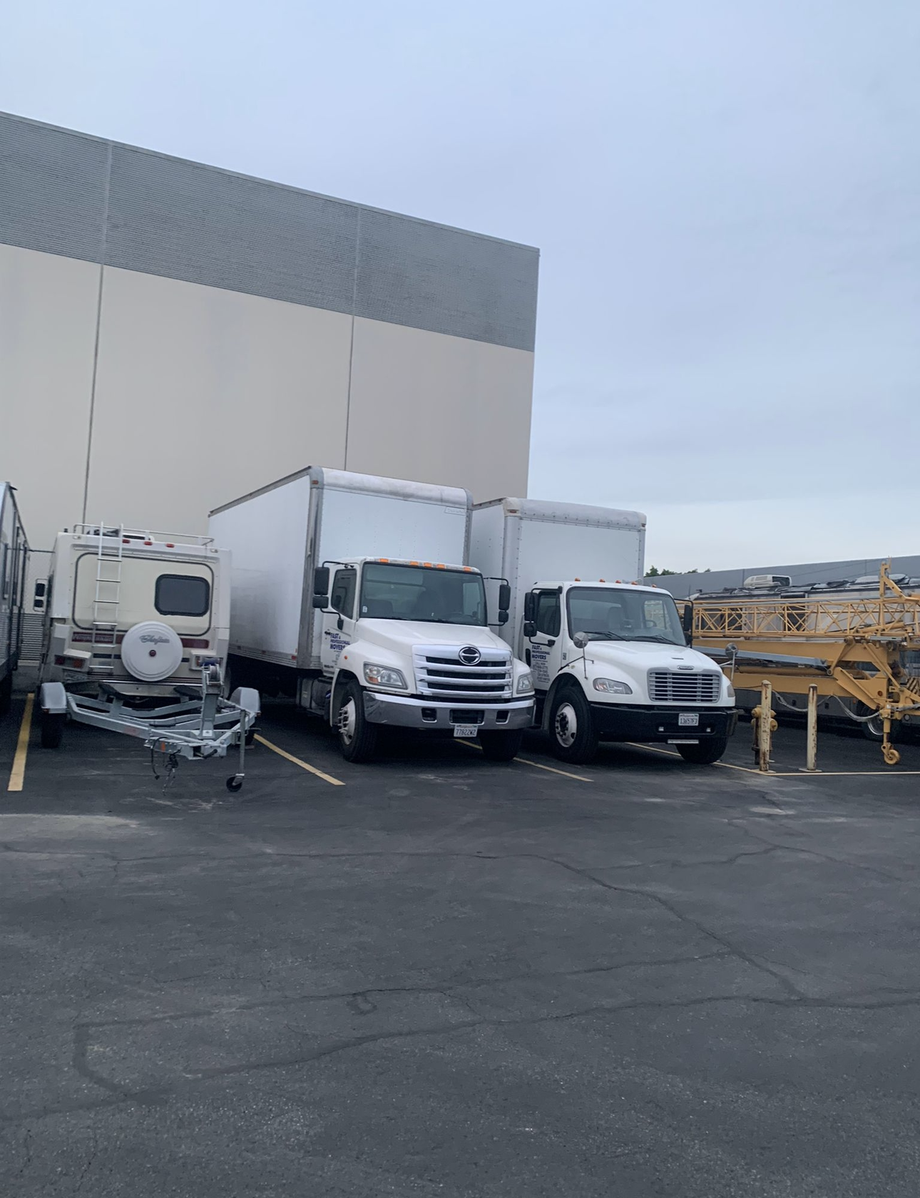 Two white box trucks parked next to a large building, a trailer is on the left side.