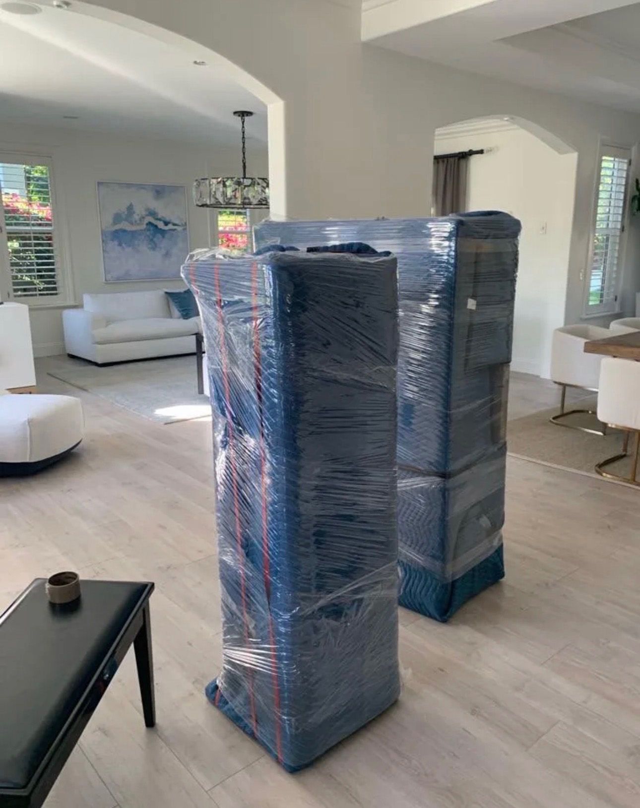 Two large, wrapped furniture items in a bright living room; white walls, light wood floors, and sofa are visible.