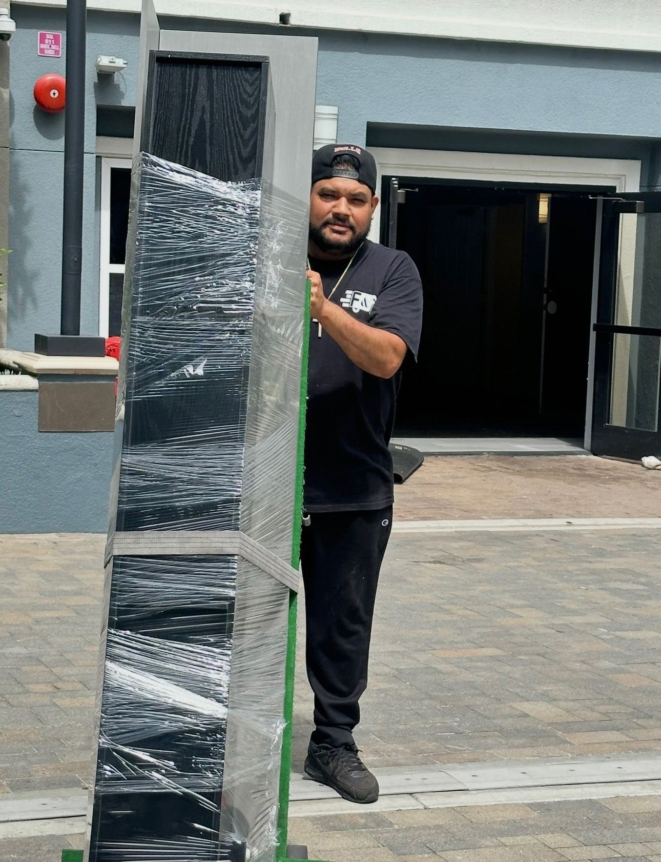 Man stands beside a tall, wrapped, rectangular object, possibly a piece of art, outside a building with open doors.