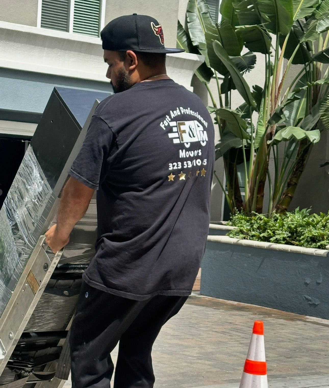 Man in black shirt with logo on back carrying a large object, likely furniture, outdoors.