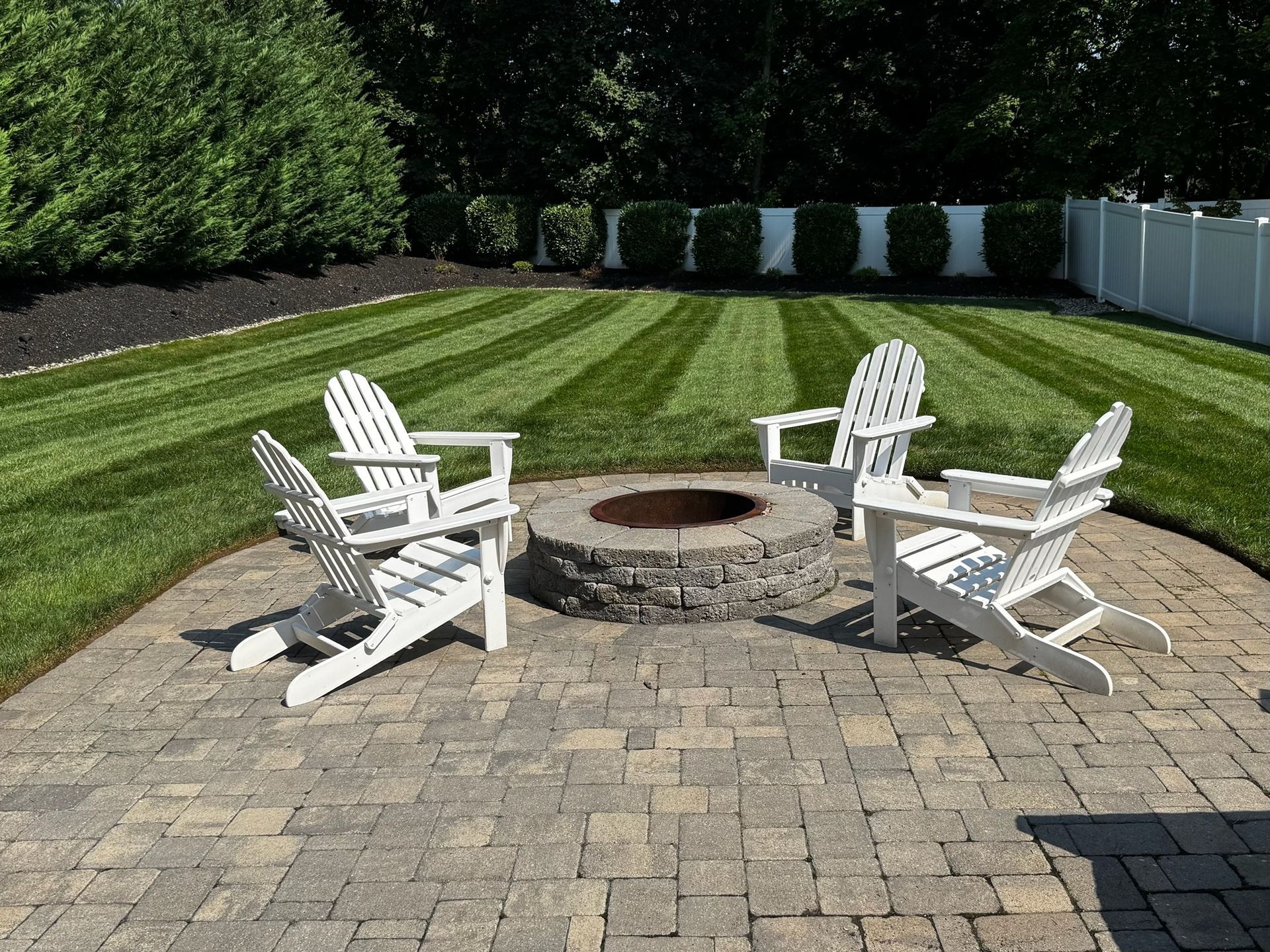 Four white Adirondack chairs around a stone fire pit on a brick patio, with a striped lawn in the background.