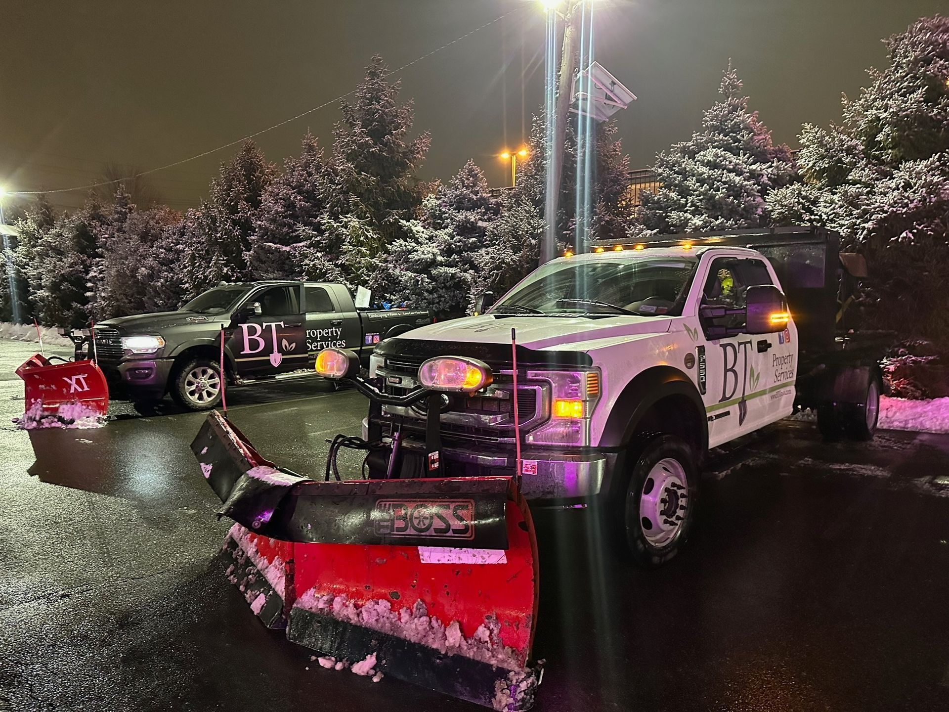 Two snowplow trucks with plows attached in a snowy parking lot at night.