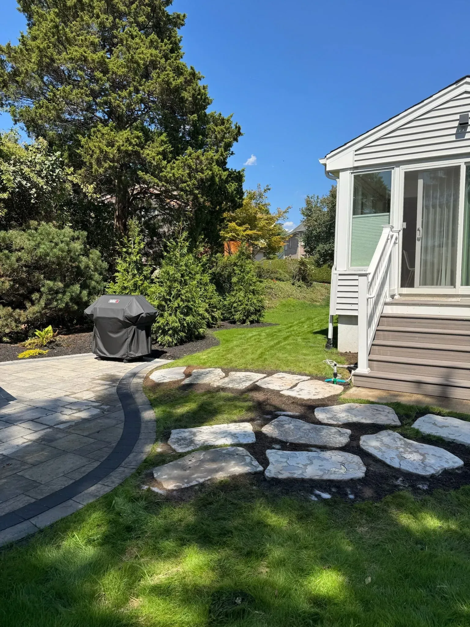 Backyard with a grill, stone path, grass, and a deck with stairs on a sunny day.