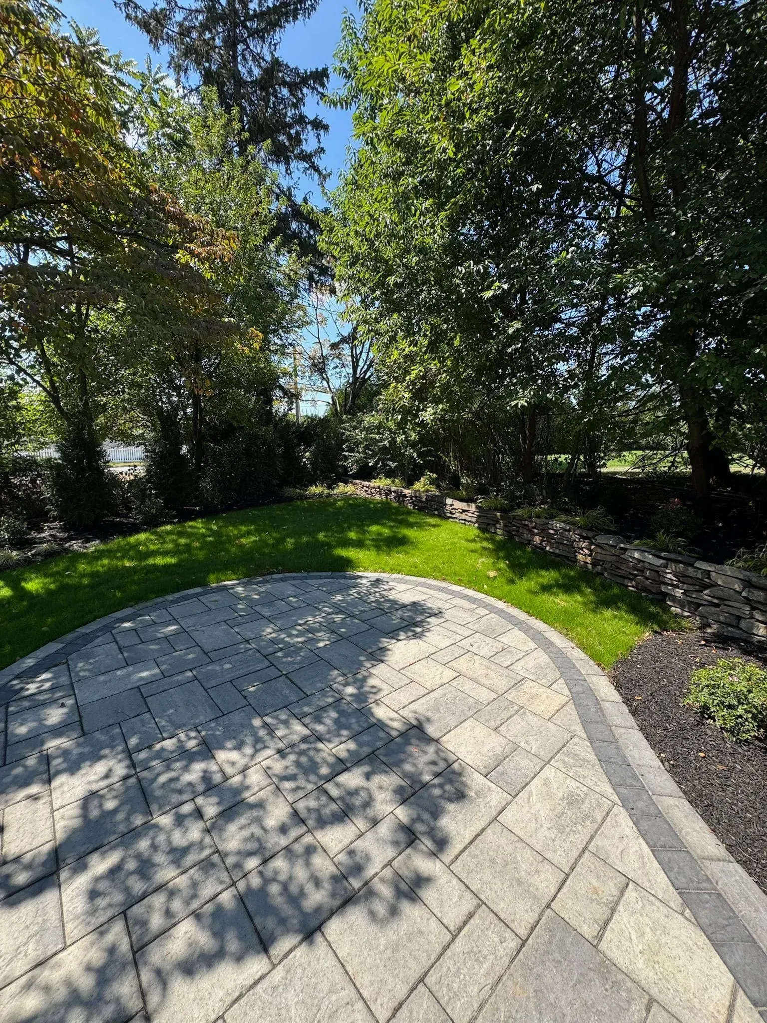 Stone patio with curved edge next to a green lawn, edged with dark mulch, under trees.