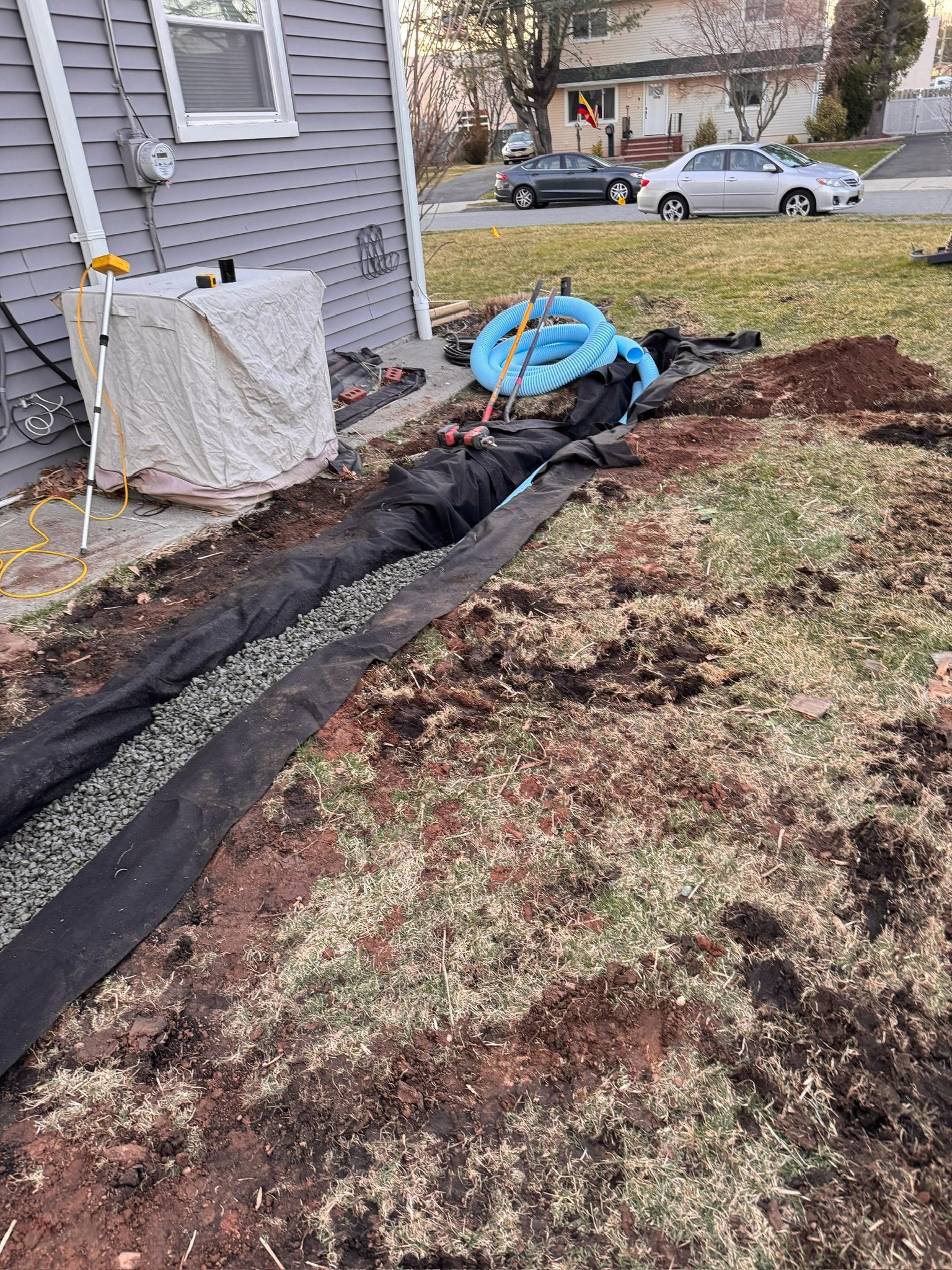 Trench with gravel and fabric being installed along a house's foundation for drainage, with blue piping nearby.