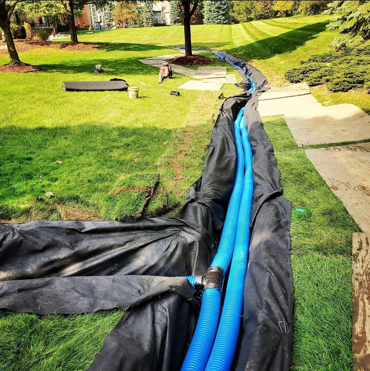 Construction site: blue drainage pipes in a trench, covered by black fabric, in a grassy yard.