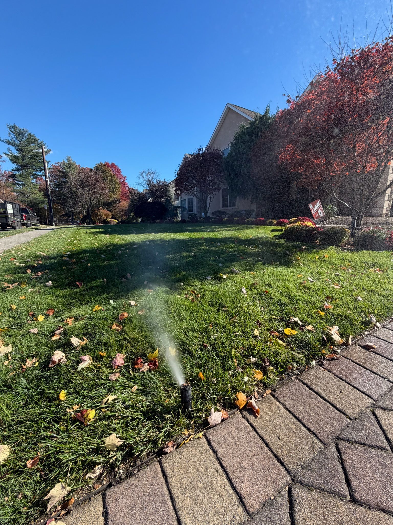 Sprinkler watering a green lawn with fall leaves, near brick walkway and trees under a blue sky.