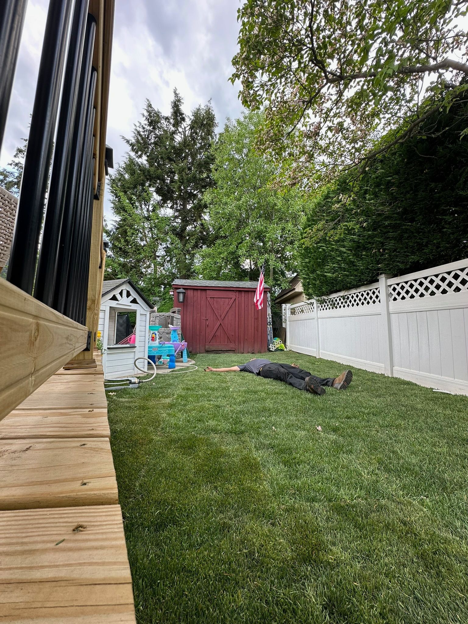 Backyard with playhouse, shed, green grass, and person laying down. White fence on the right. Deck on the left.
