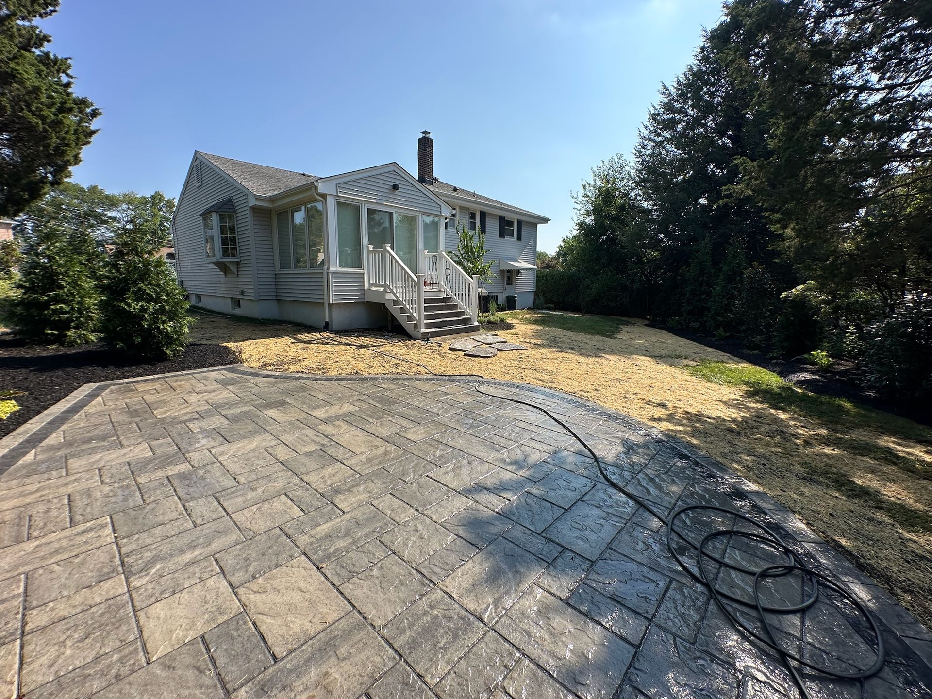 Stone patio in front of a house, leading to stairs. Gravel and trees surround the home.