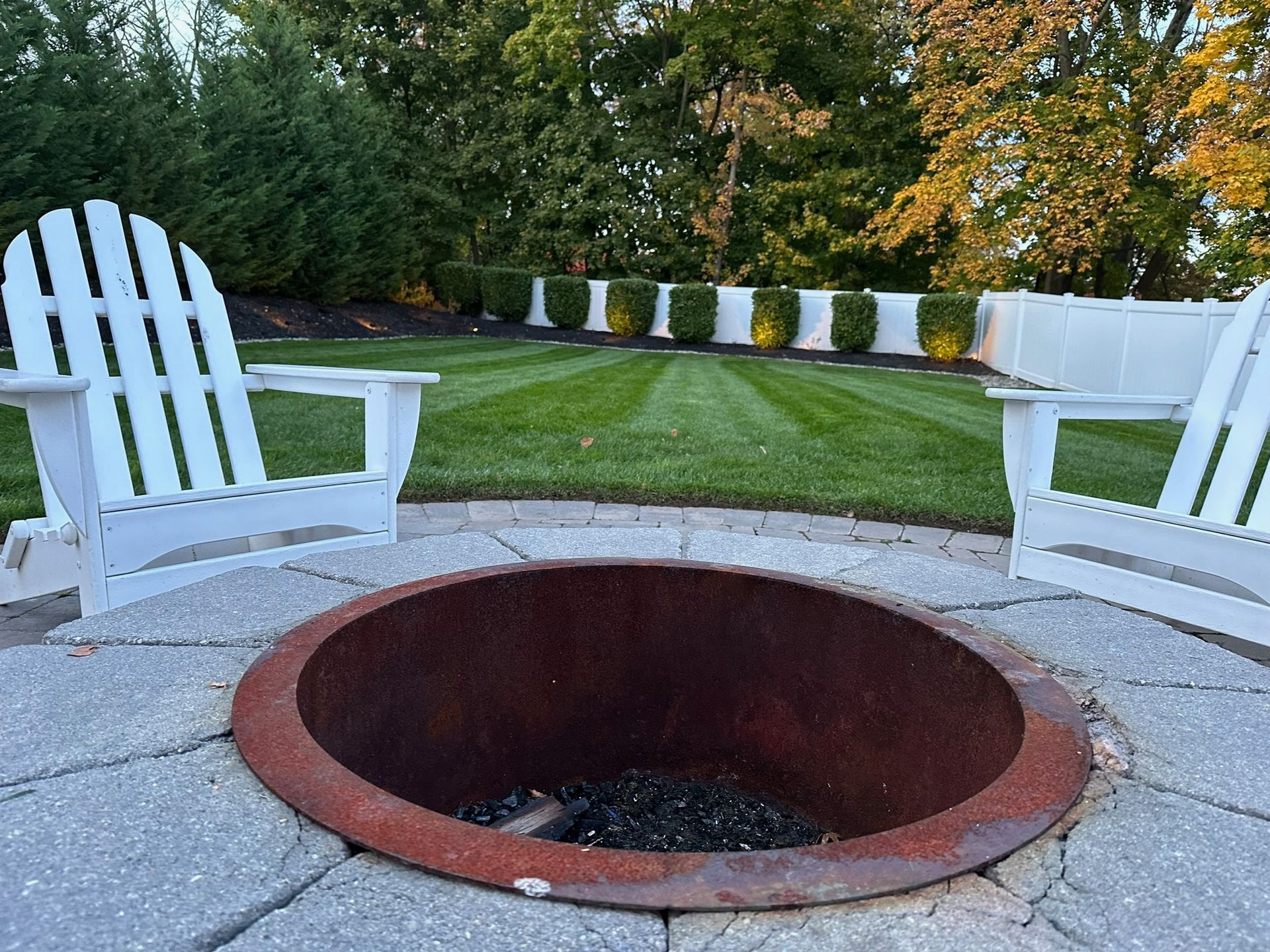 Two white Adirondack chairs face a rustic fire pit on a stone patio, with a manicured lawn and green hedge in the background.