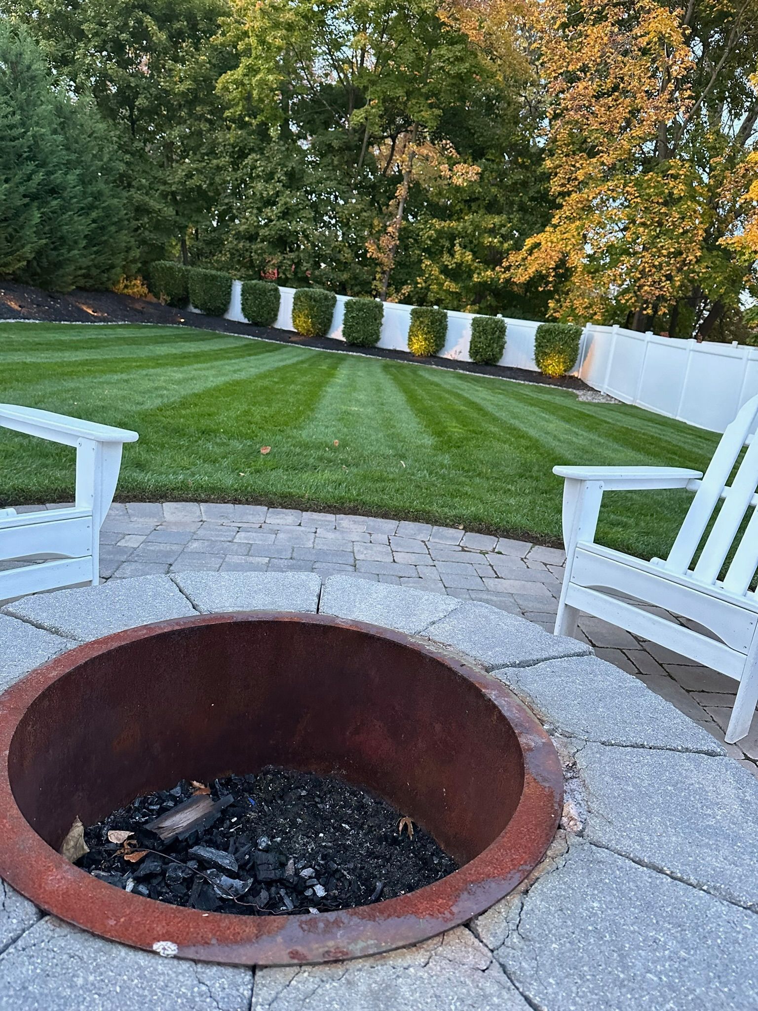 Fire pit on a stone patio with white chairs, looking out over a mowed lawn with trees and a white fence.