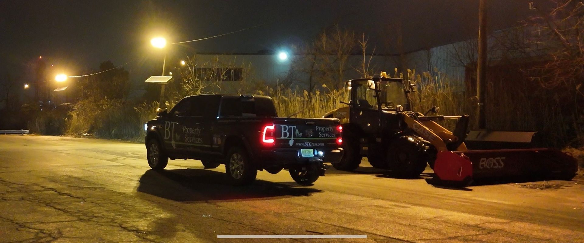 A dark pickup truck towing a plow on a snowy road at night, illuminated by streetlights.