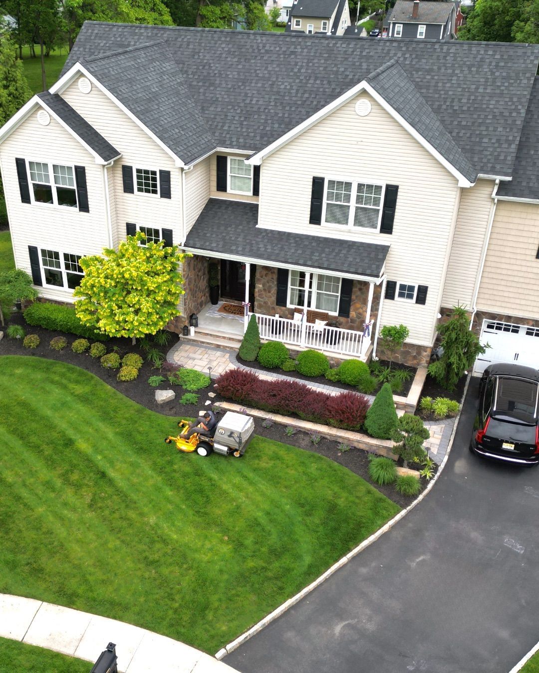 Two-story house with black roof, manicured lawn, and parked car; lawnmower in the yard.