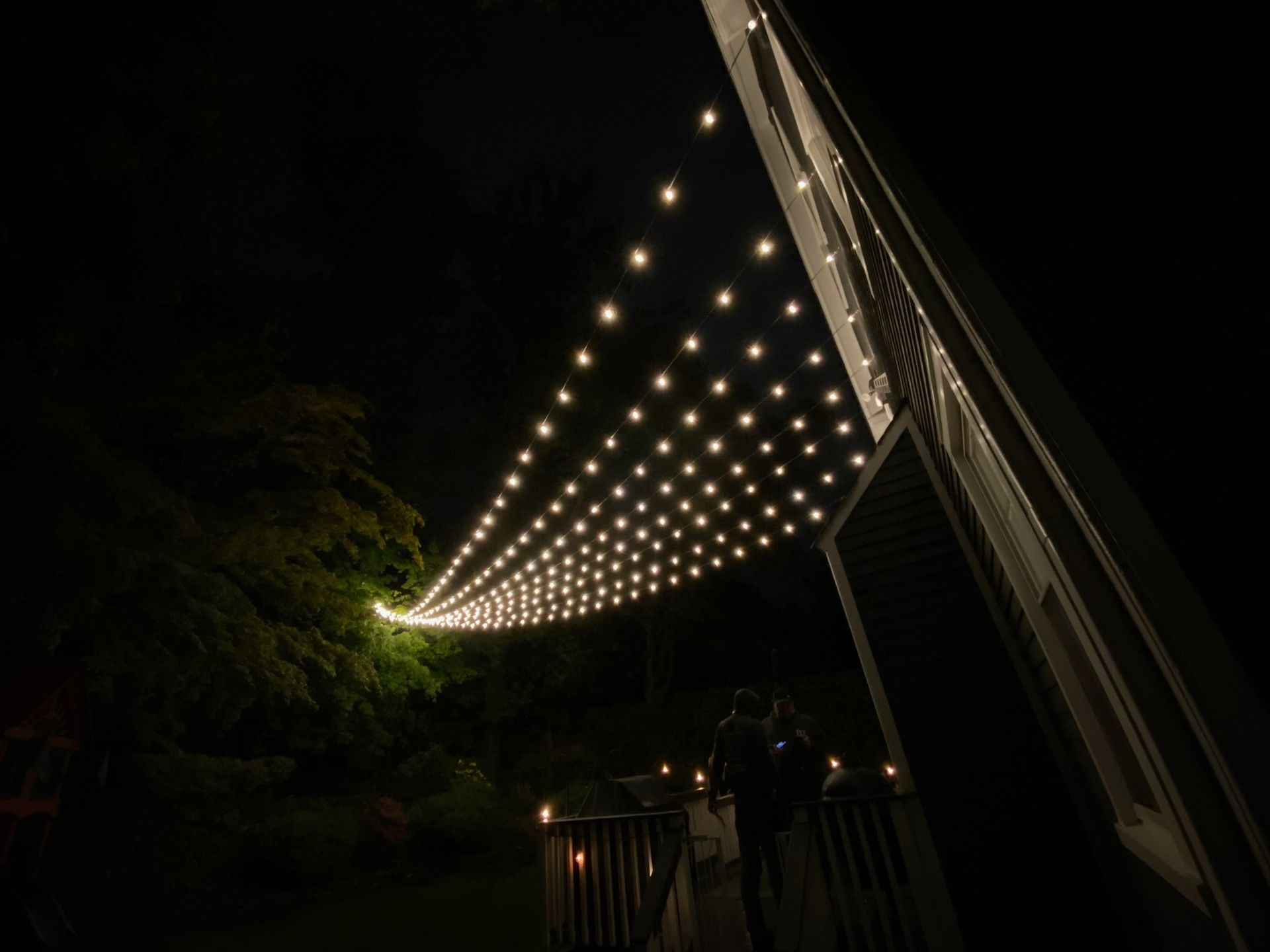 String lights illuminate a dark night. The lights are strung across a deck, connected to a building on the right.