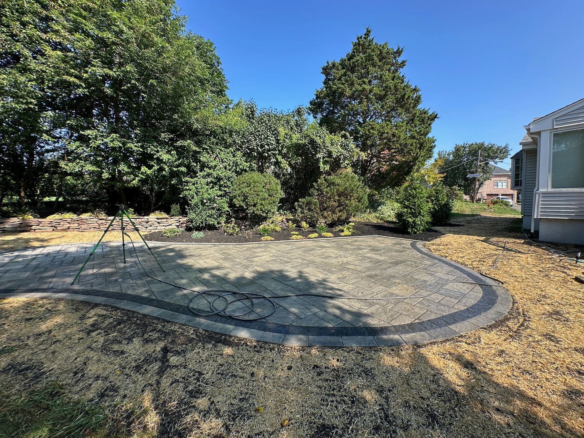 Gravel patio edged with dark pavers, surrounded by grass and shrubs on a sunny day.