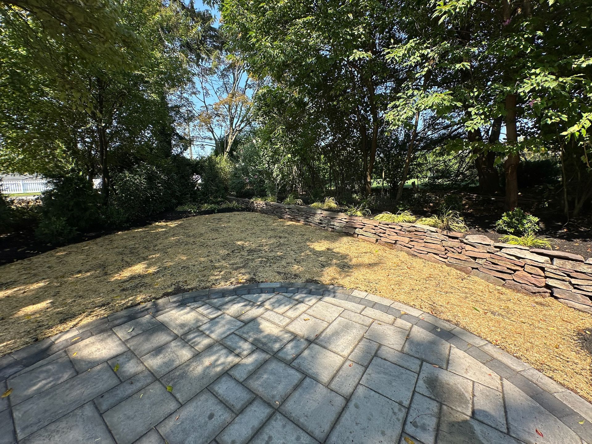 A paved patio transitions to a gravel area, surrounded by a stone wall and lush green trees.