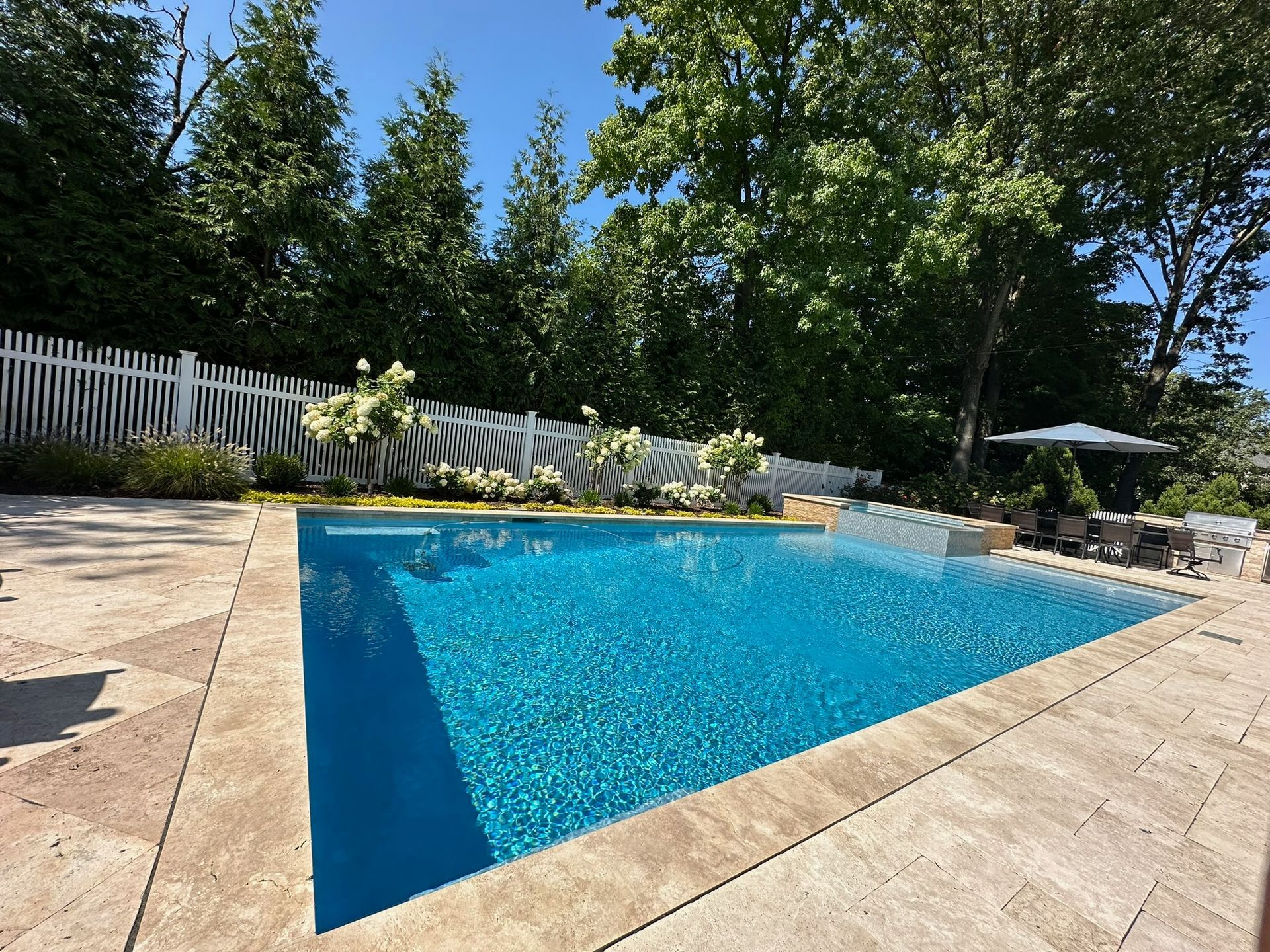 A rectangular swimming pool with blue water surrounded by beige tiles, a white fence, and trees.