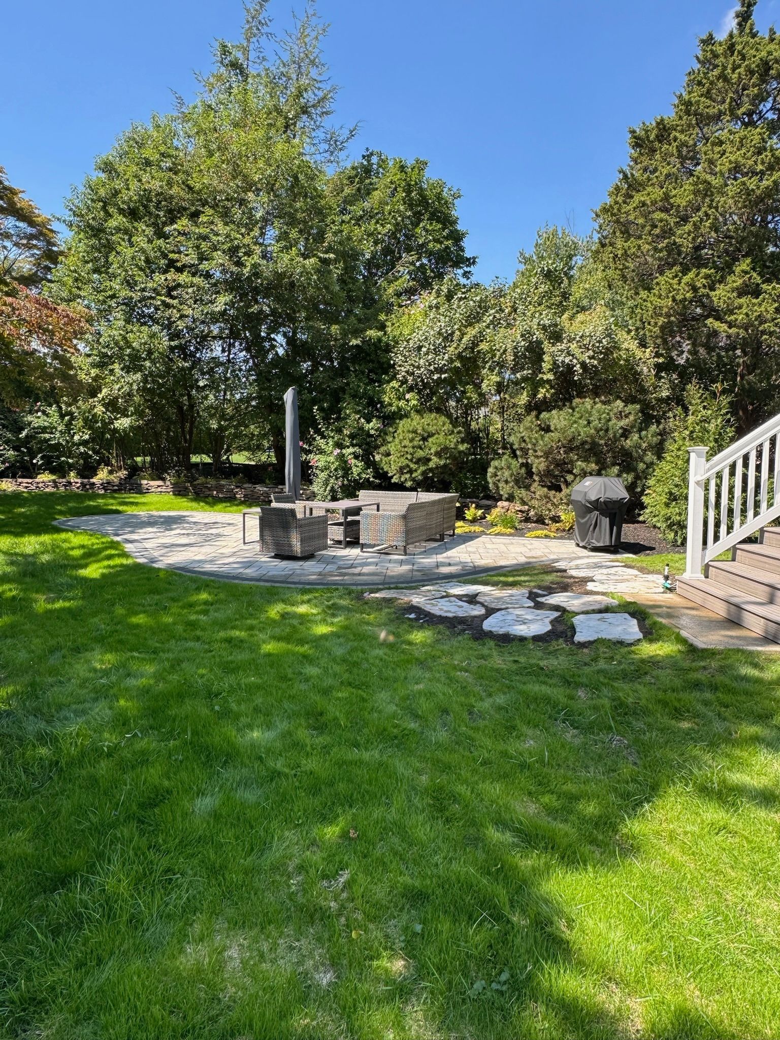 Lawn with stone patio and furniture, surrounded by trees. Sunny day with a white deck railing on the right.