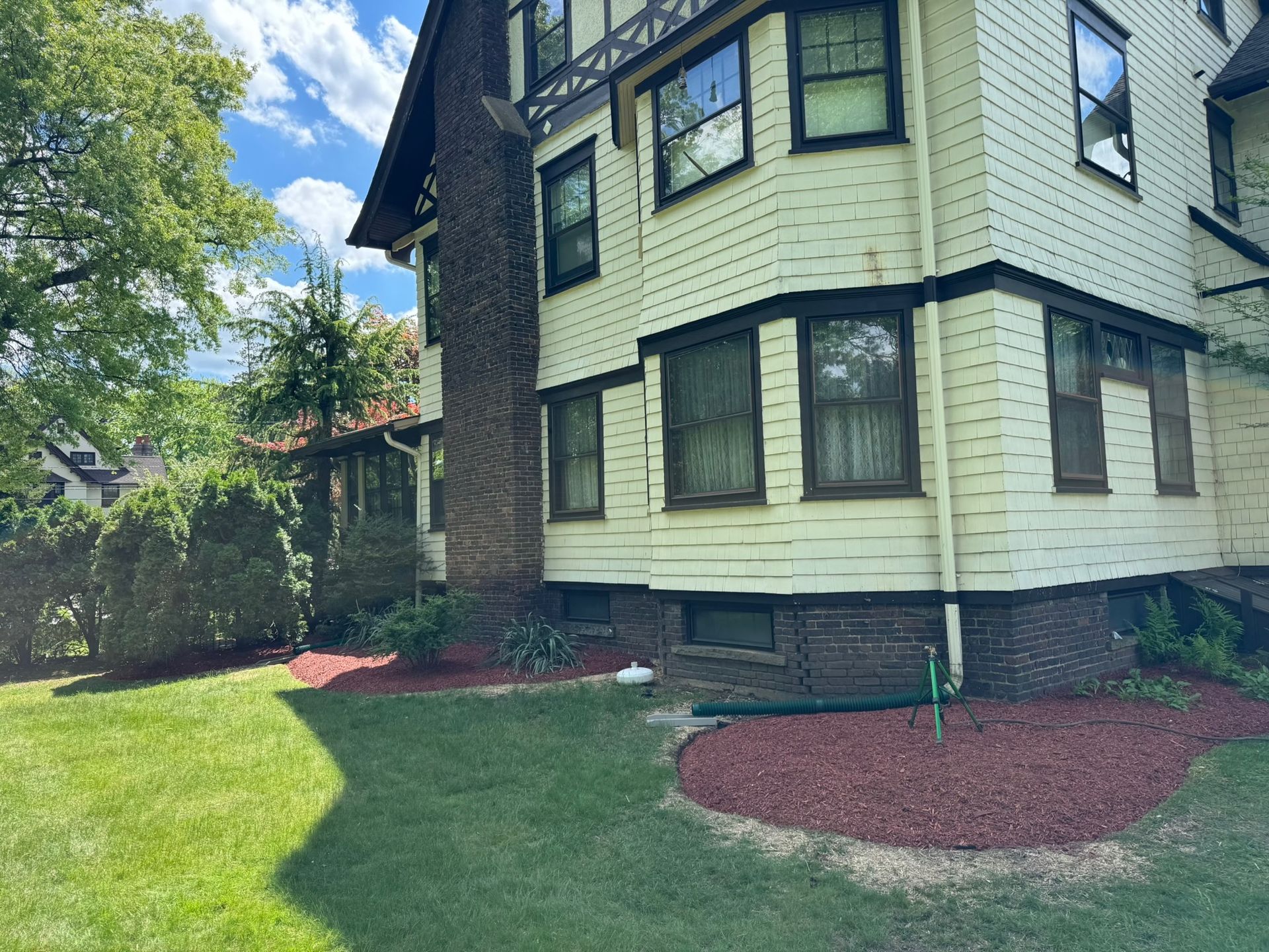 Side view of a light yellow house with dark trim, chimney, and red mulch landscaping.