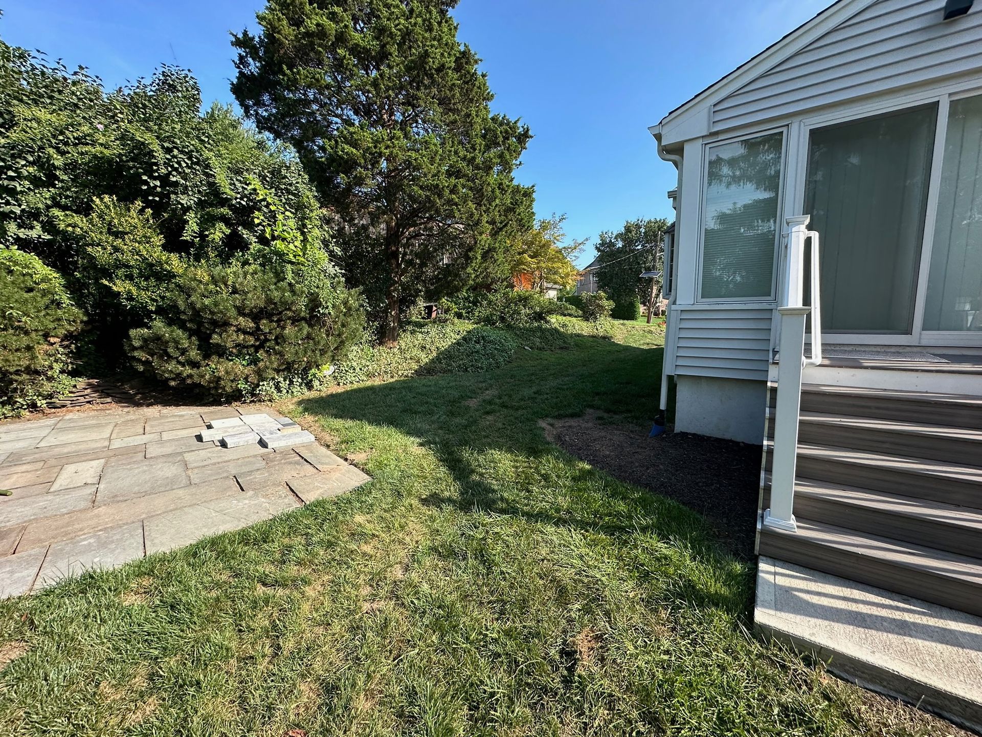 Grassy backyard with a stone patio, shrubs, and a house with a deck.
