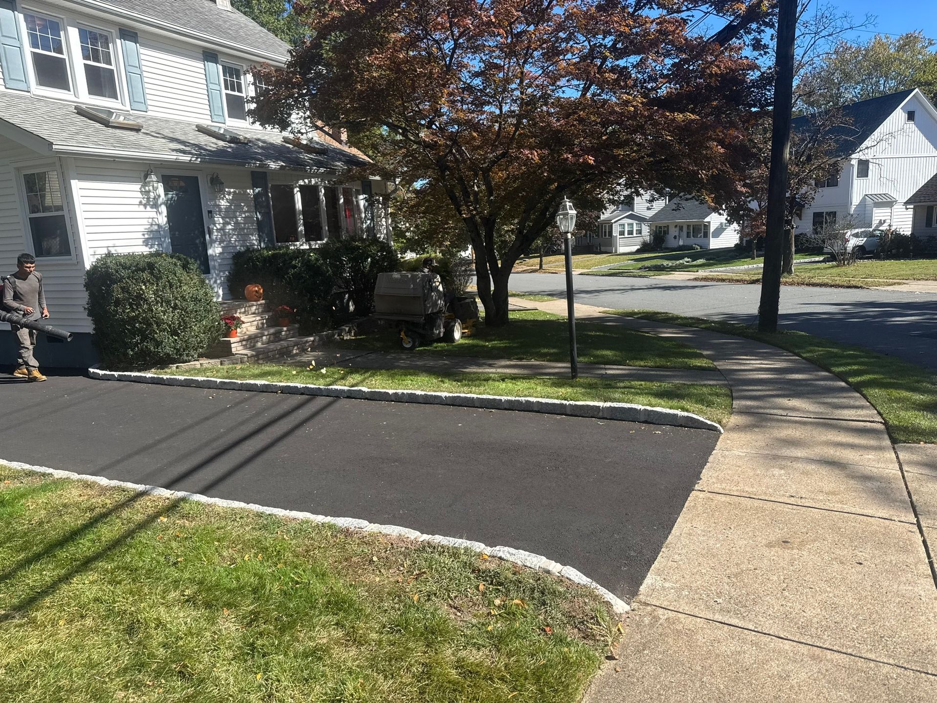 Man blowing leaves near a newly paved asphalt driveway and sidewalk in front of a house.