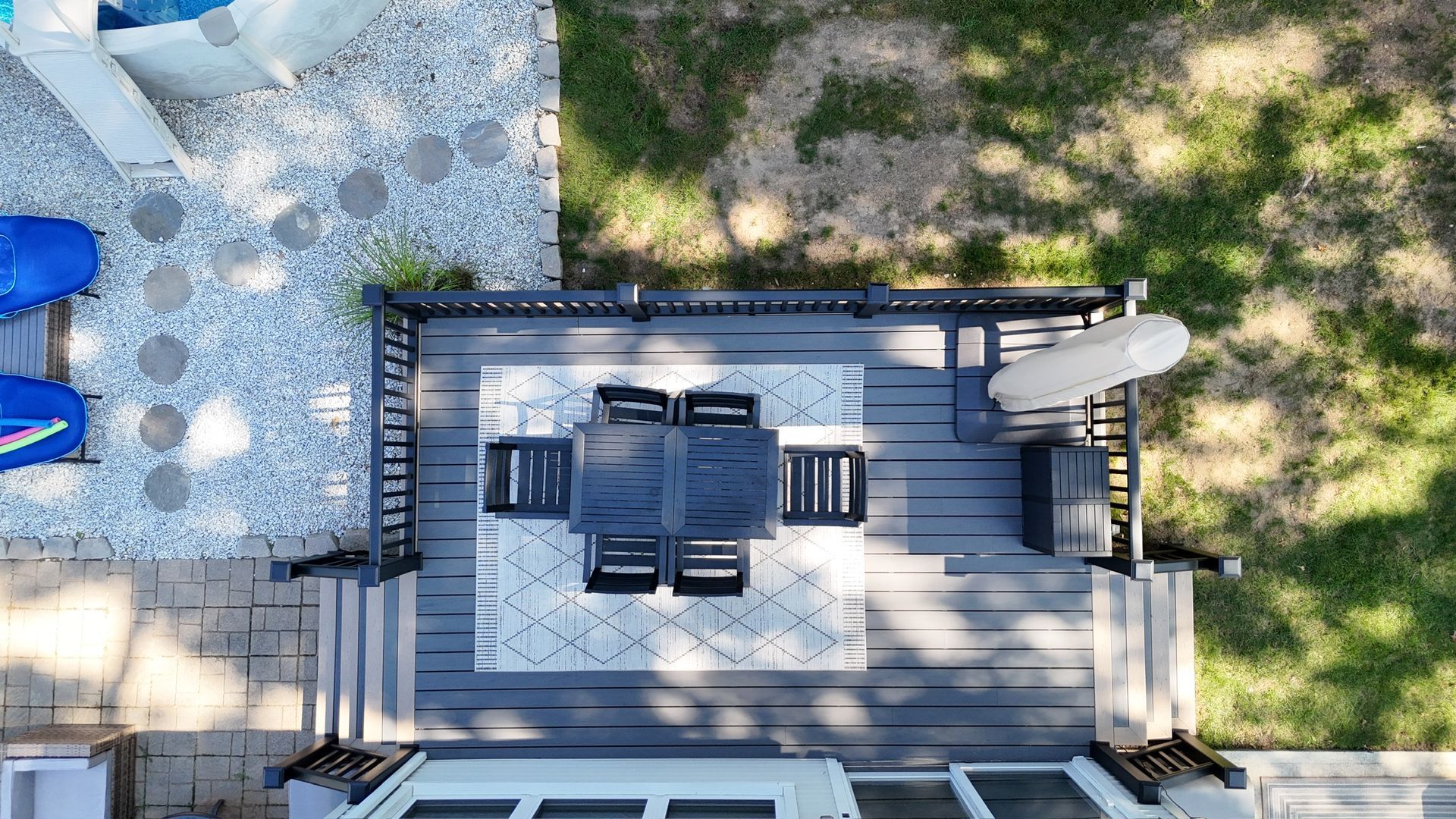 Overhead view of a deck with a table, chairs, and umbrella. A pool and lawn are nearby.