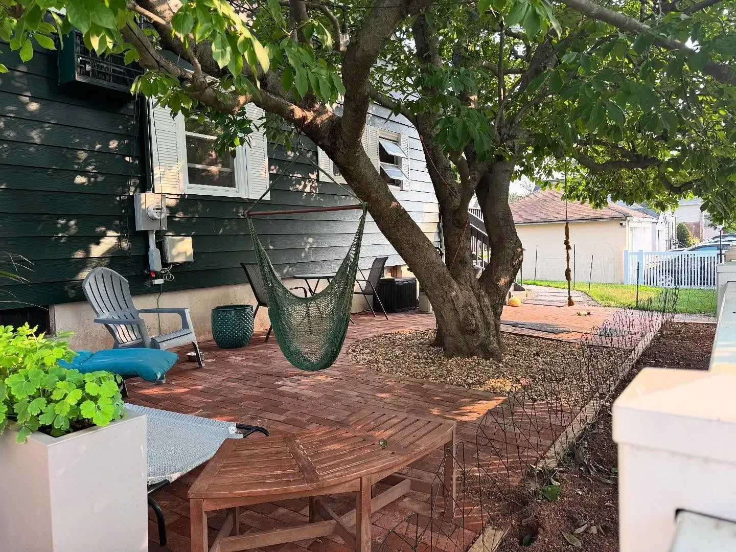 A backyard patio with a tree, chair, and hammock. Red brick and gravel surround. Green plants in a planter.