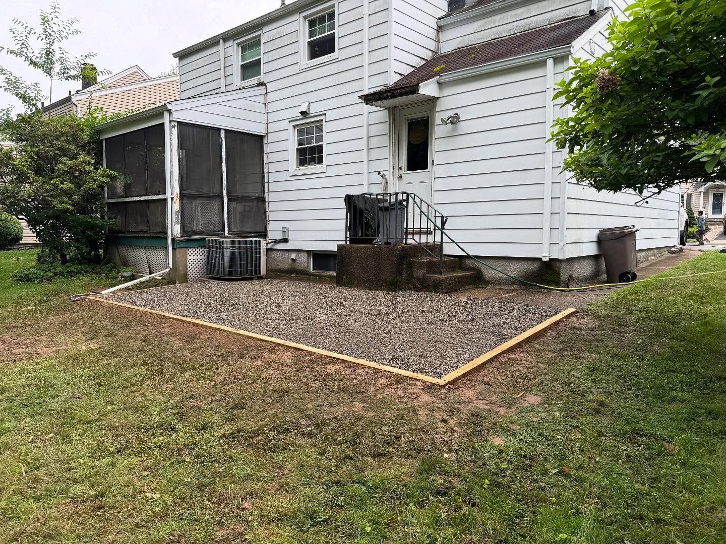 Gravel patio with wooden border next to a white house with a screened porch and green grass.