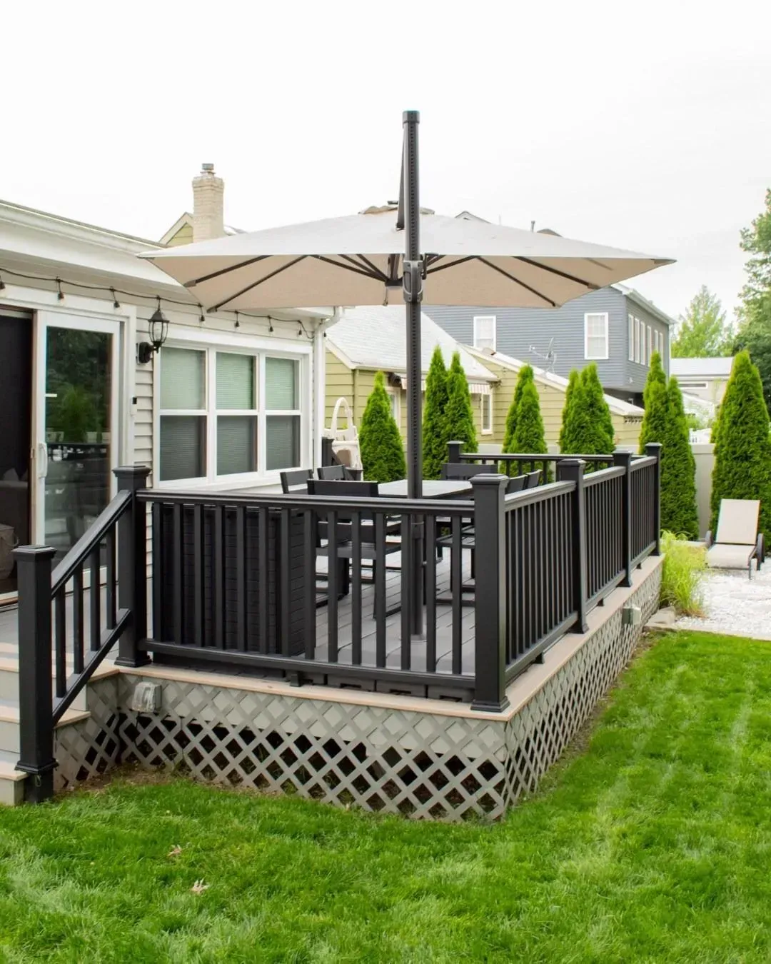 Black deck with a tan umbrella, surrounded by green grass and landscaping, next to a house.