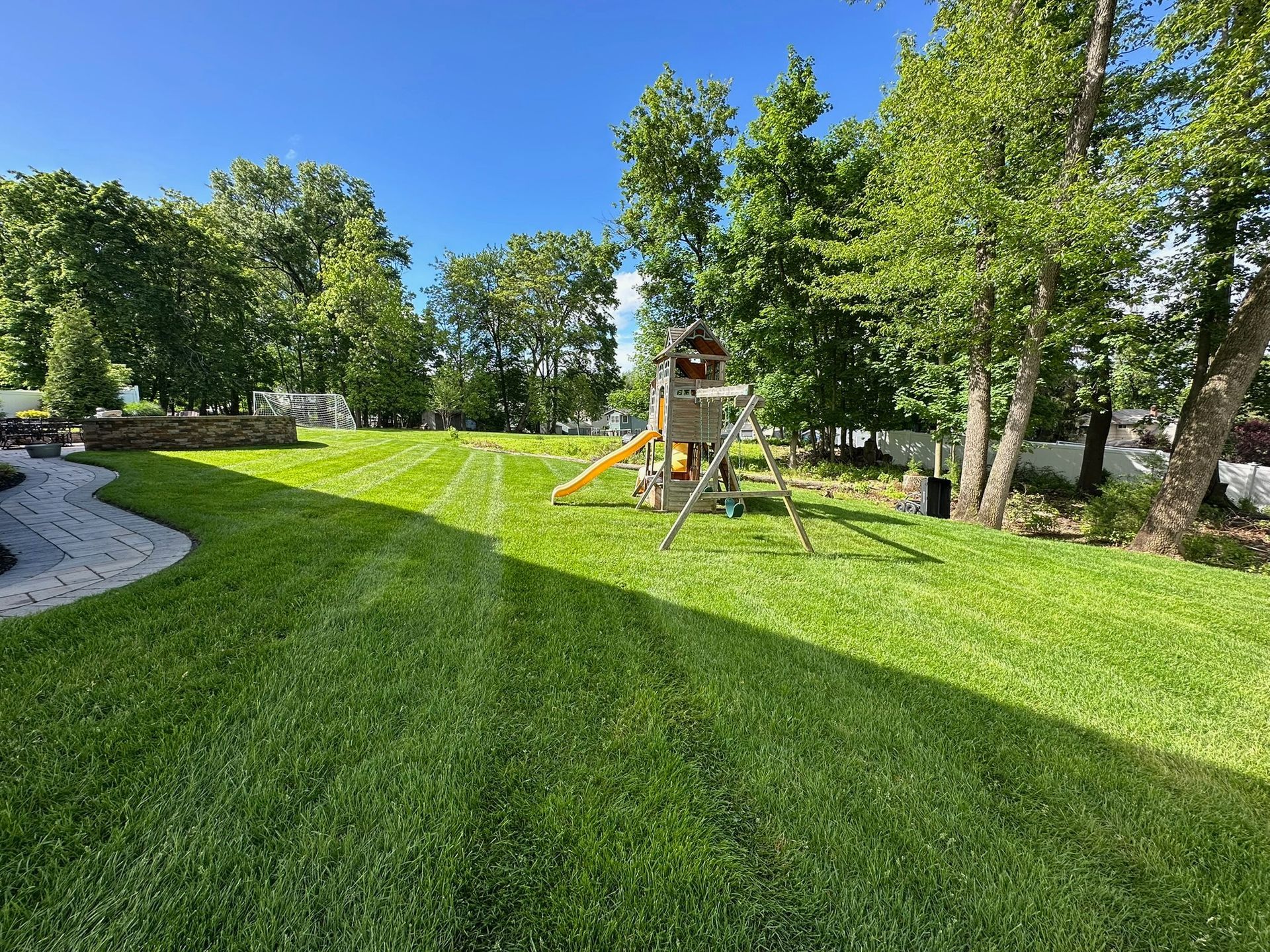 Lush green backyard with freshly mowed grass. A wooden playset sits near the trees under a bright blue sky.