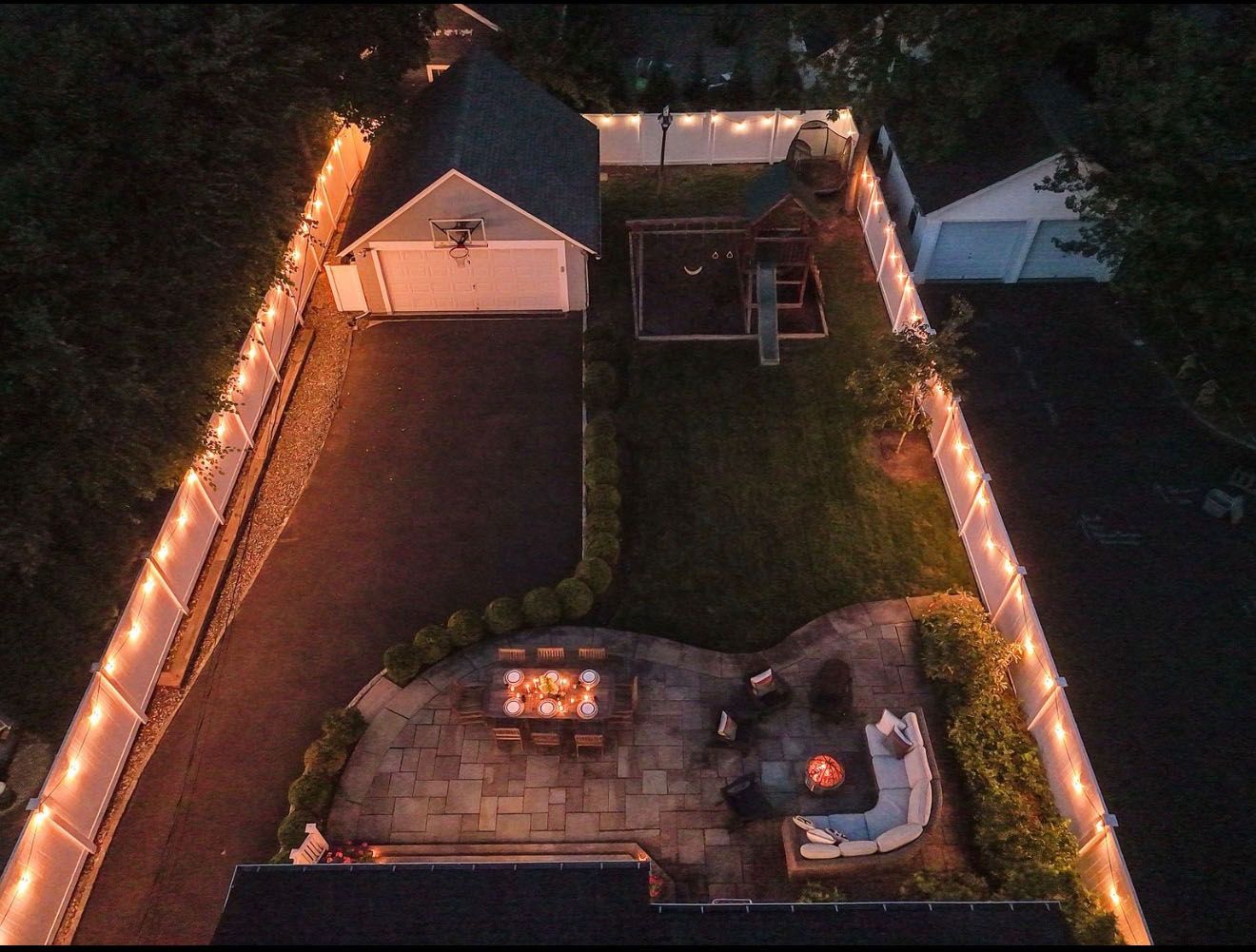 Aerial view of a backyard with string lights on a fence, garage, playground, and patio with a fire pit.