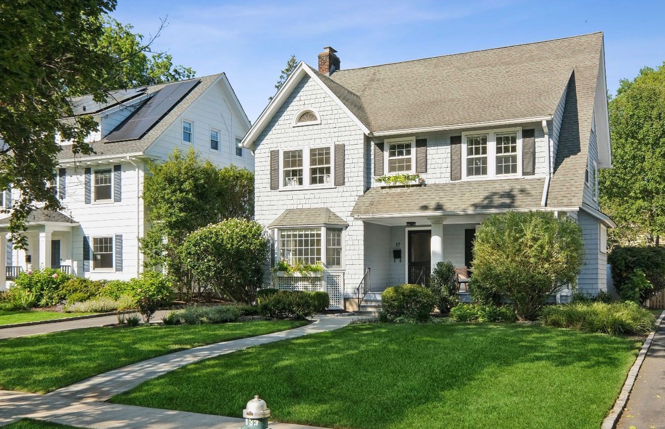 Blue and white two-story house with green lawn, trees, and small shrubs in a suburban neighborhood.