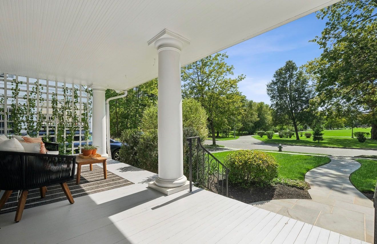 White porch with a column, wicker furniture, and a view of a green landscape and a road.