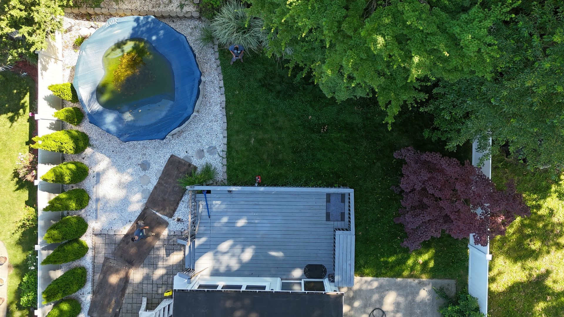 Aerial view of a backyard with a round pond, stone patio, and deck beside green lawn and trees