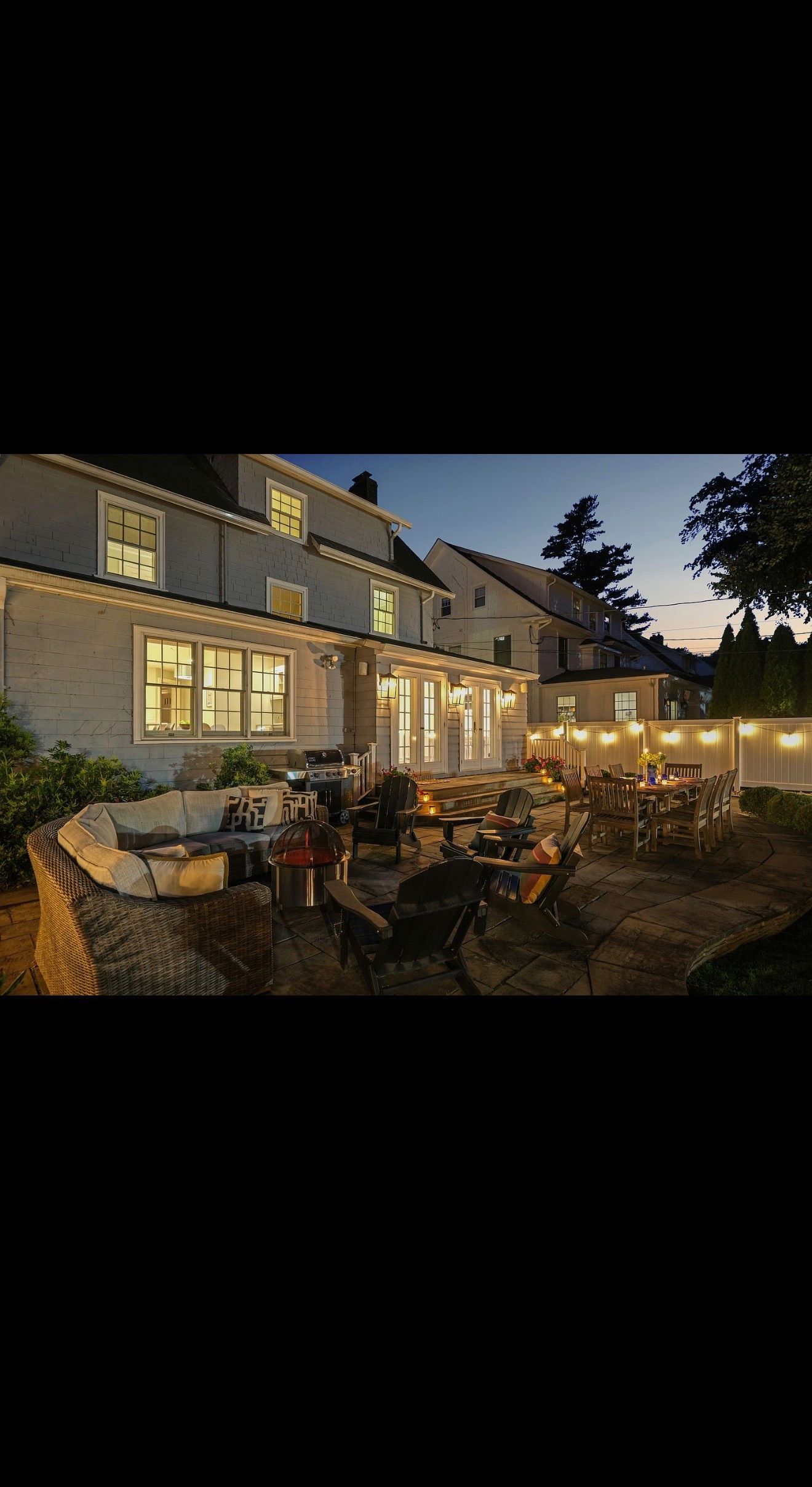 Lit stone house with glowing windows and a patio, surrounded by outdoor tables at dusk