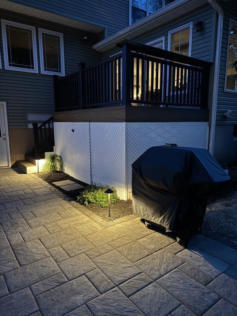 Lit backyard patio with deck railing, stone pavers, and a covered grill at dusk