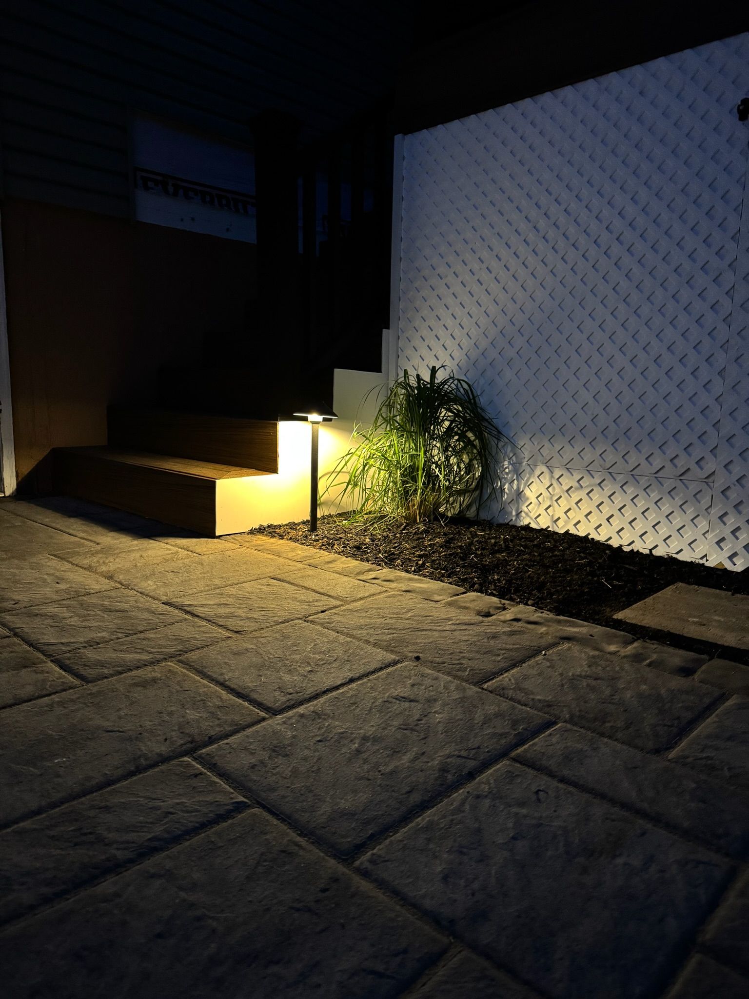 Dimly lit stone patio with a glowing step light, potted plant, and white textured wall at night