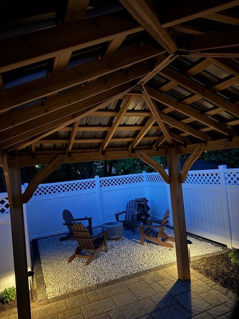 Covered patio with wooden roof, patio chairs, fire pit, and lit gravel yard beside a white fence.