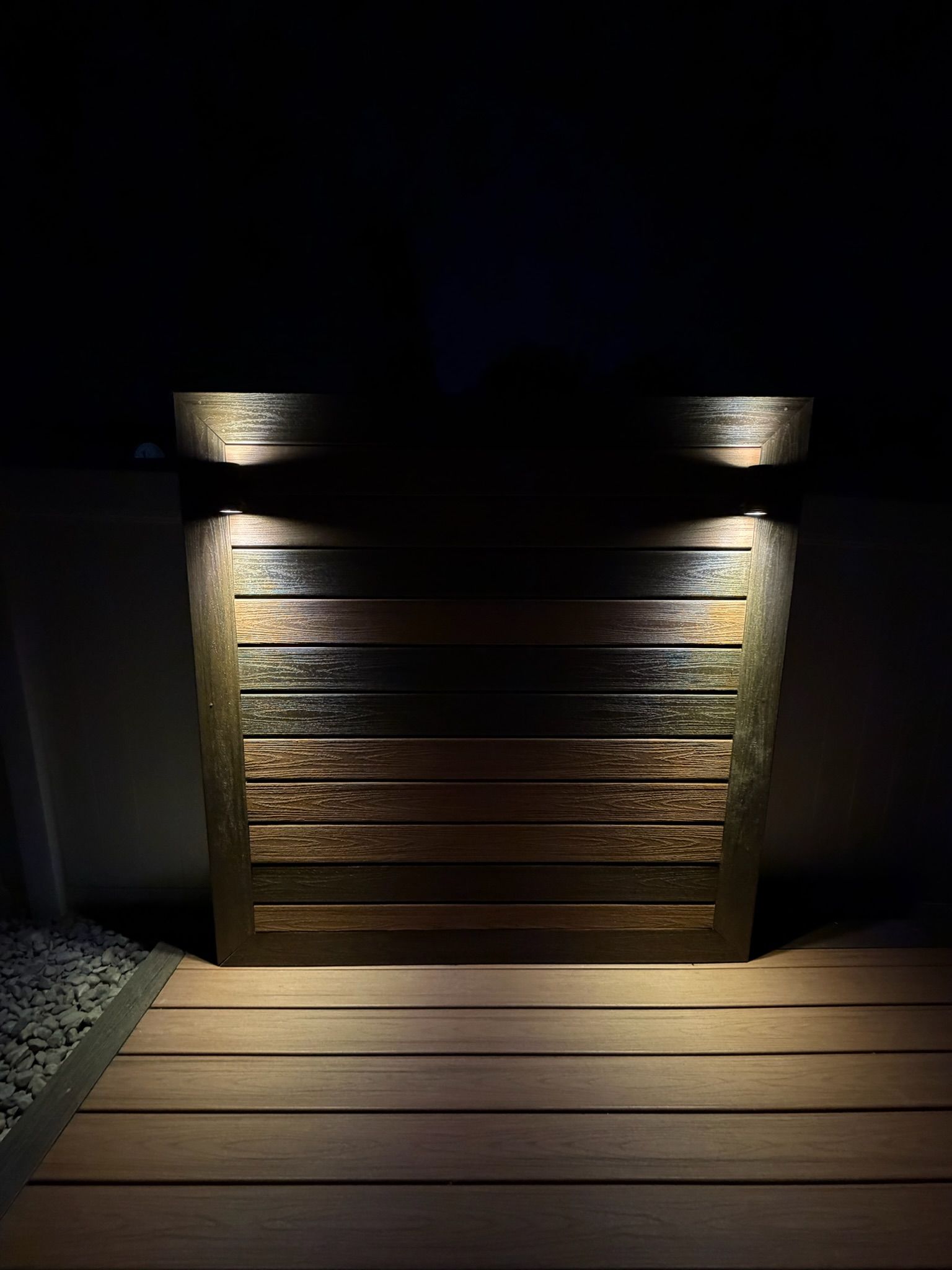 Dimly lit wooden planter or wall with two small lights above, next to a wooden deck at night