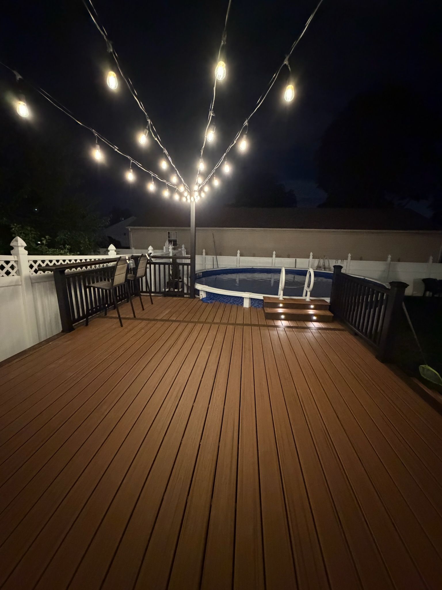 String lights over a wooden deck at night with a small above-ground pool and white fence.