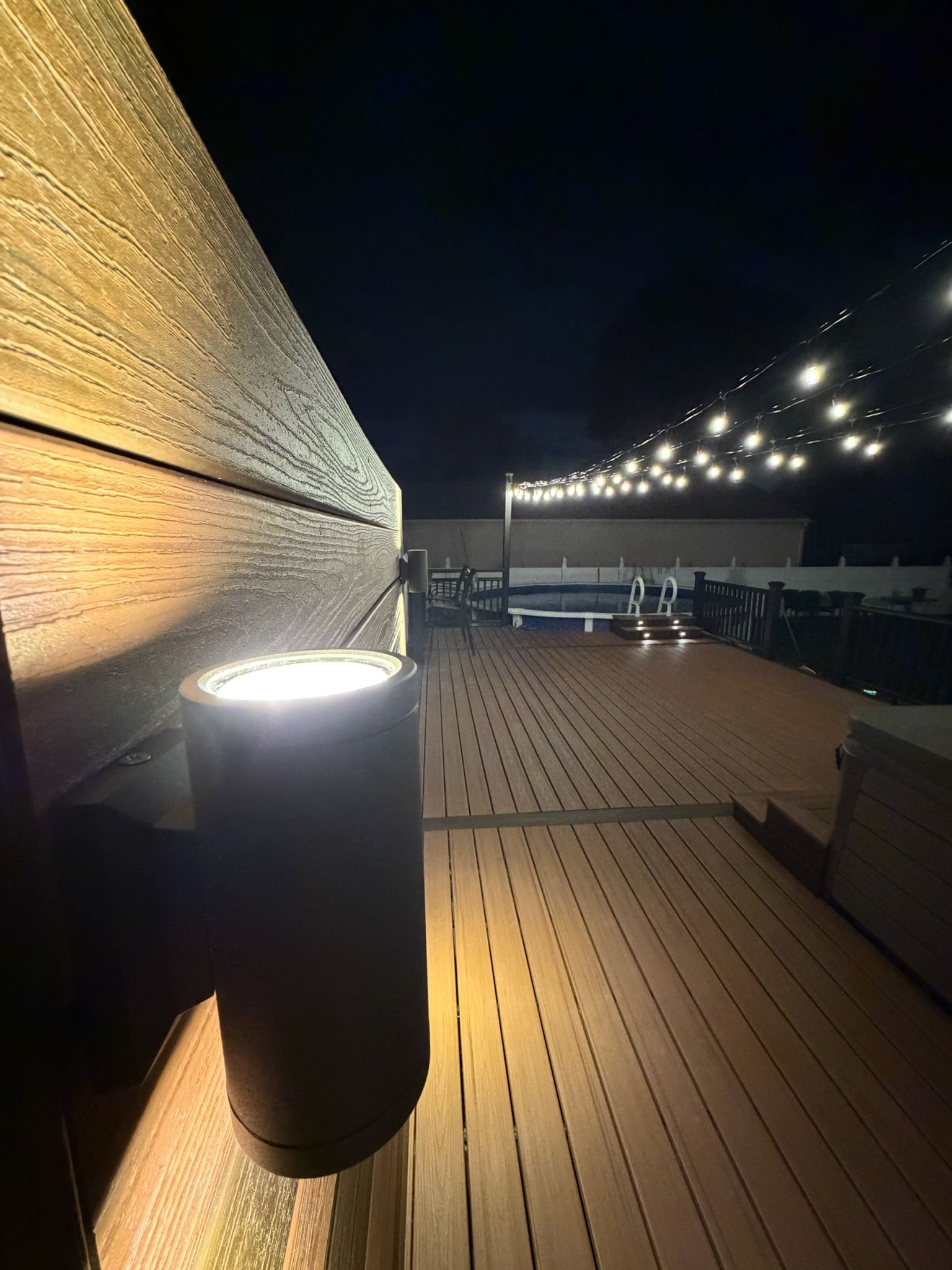 Wooden deck at night with string lights, a lit wall, and a cylindrical lamp on the left.