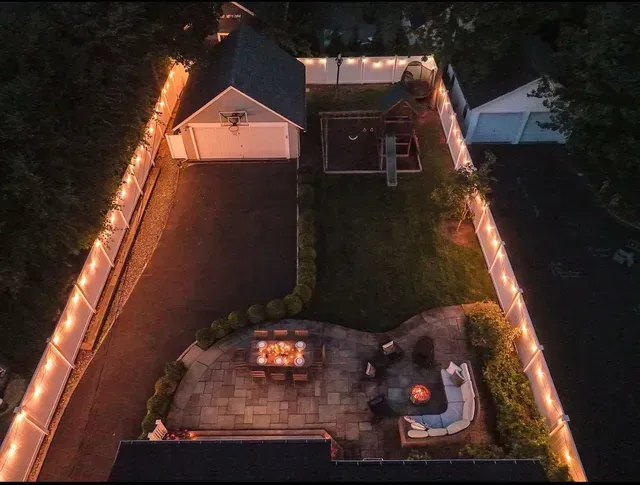 Aerial view of a backyard at night with string-lit fence, patio fire pit, and a small pool or hot tub.