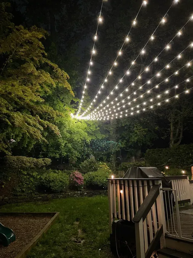 Backyard garden at night with string lights overhead and a wooden deck railing in the foreground