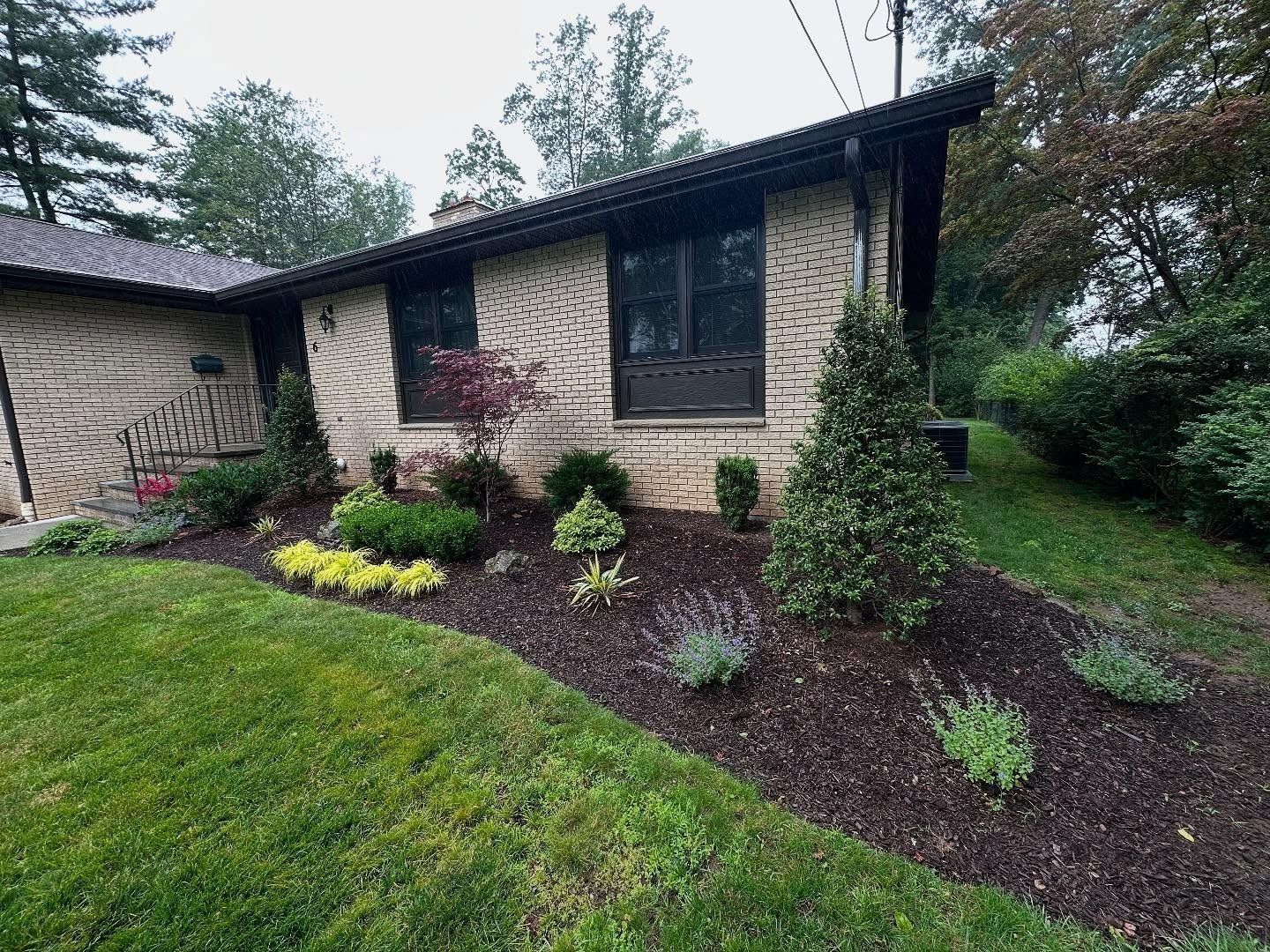 Landscaped brick house with dark roof, mulched garden beds, and green lawn on a rainy day