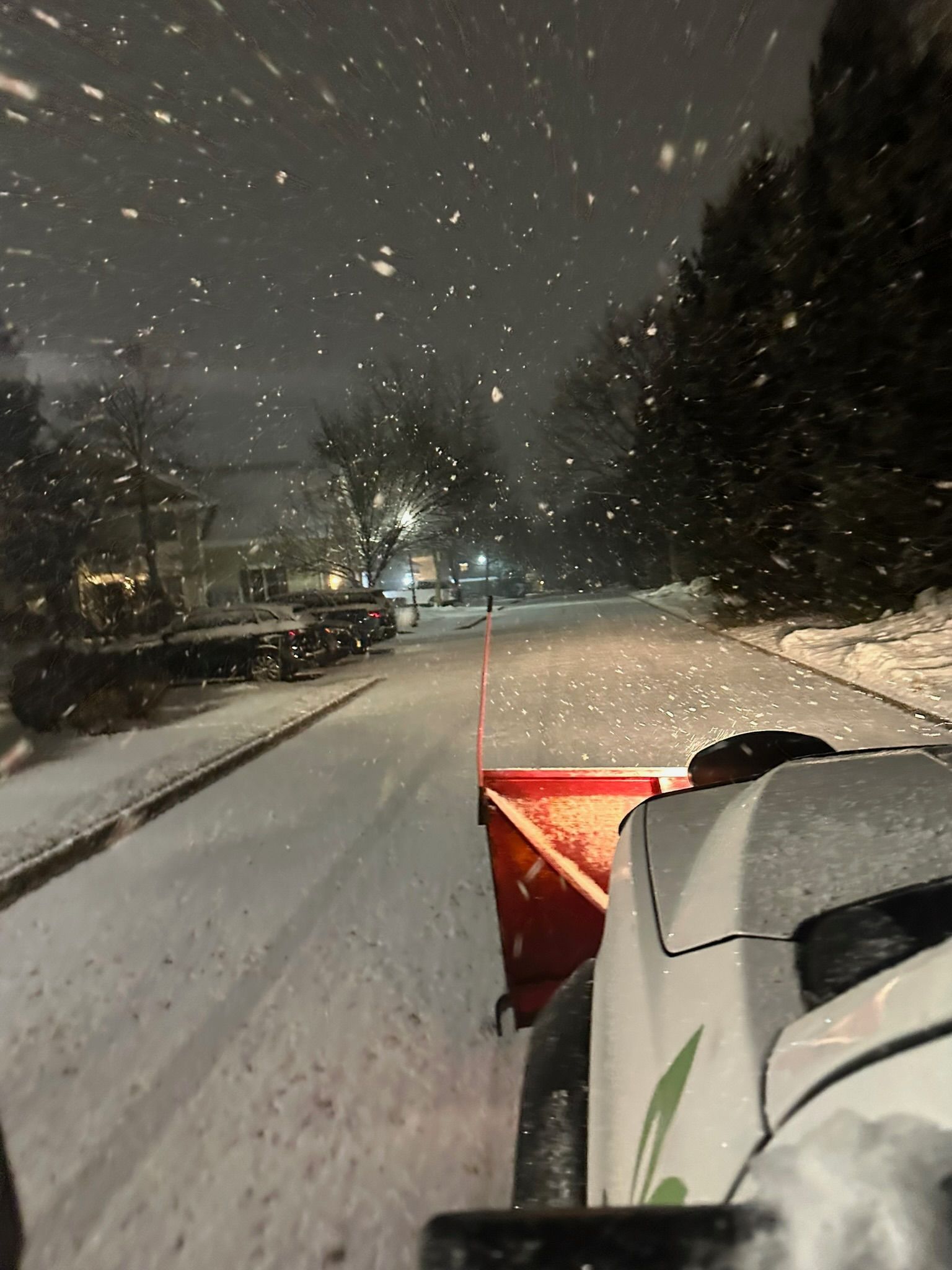 Snowplow clearing a snowy residential street at night, with snow falling and parked cars along the road